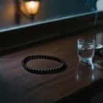 Evening Ritual by Yuano: A sacred obsidian necklace resting on a wooden altar beside a glass of water and incense at dusk.