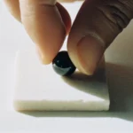 The First Stone by Yuano: A sacred obsidian bead being placed on a white stone base by an artisan's hand.