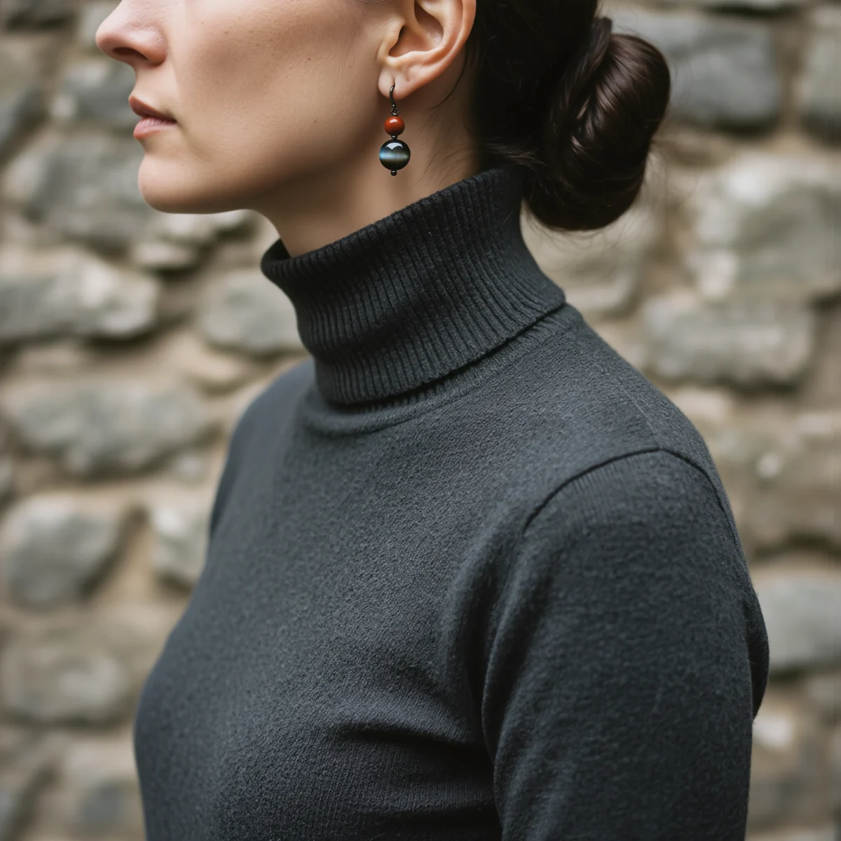 Fortress No. 10 — Lifestyle Observational Poise Fortress No. 10: Ancient Shield earrings worn by a model in a dark grey turtleneck against an ancient stone wall.