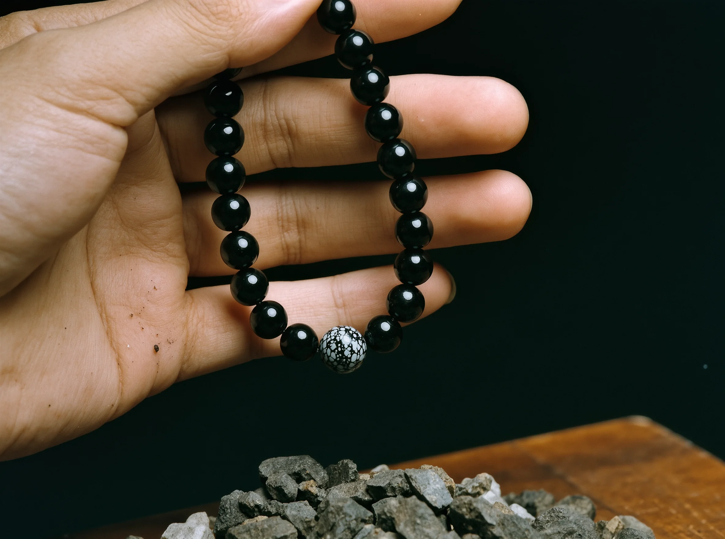 Home 5 Sovereign Stillness Jewelry by Yuano: An artisan's hand holding a polished obsidian necklace with a celestial focal bead over raw mineral fragments.